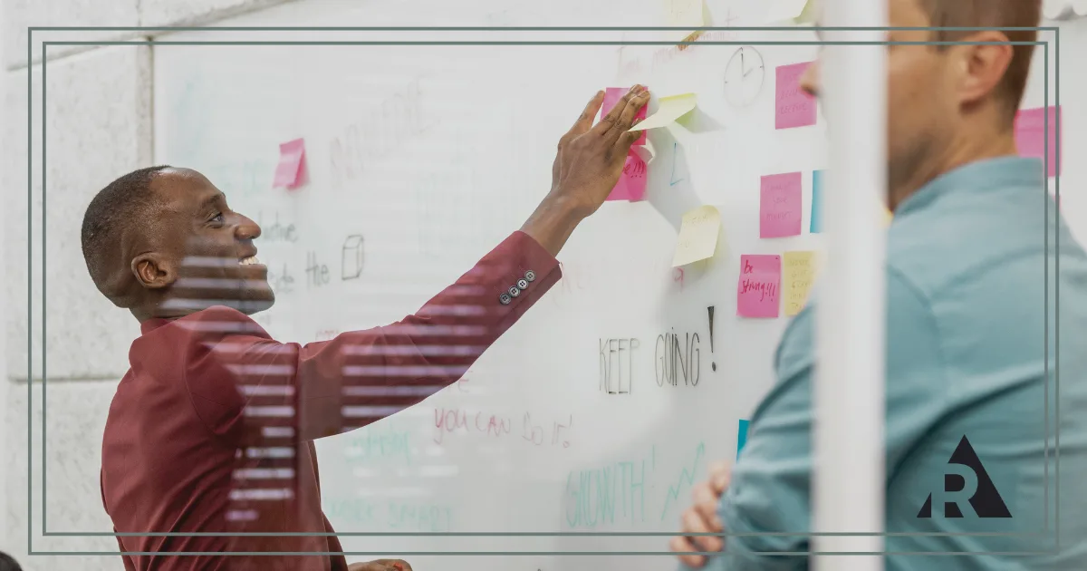 Two professionals collaborate in a meeting room, standing beside a glass whiteboard covered with sticky notes and diagrams.