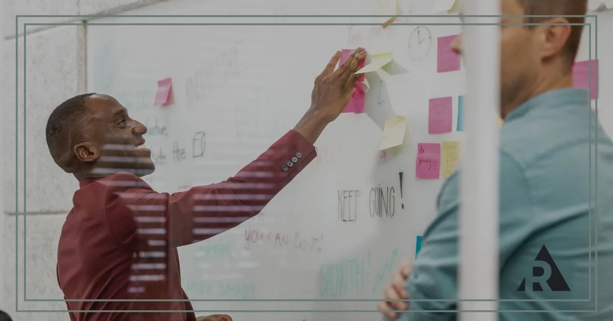 Two professionals collaborate in a meeting room, standing beside a glass whiteboard covered with sticky notes and diagrams.