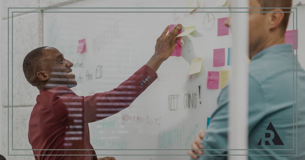 Two professionals collaborate in a meeting room, standing beside a glass whiteboard covered with sticky notes and diagrams.